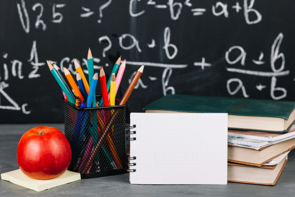 Biurko z jabłkiem, kolorowymi kredkami, otwartym zeszytem w kratkę i książkami, na tle tablicy z wzorami matematycznymi.