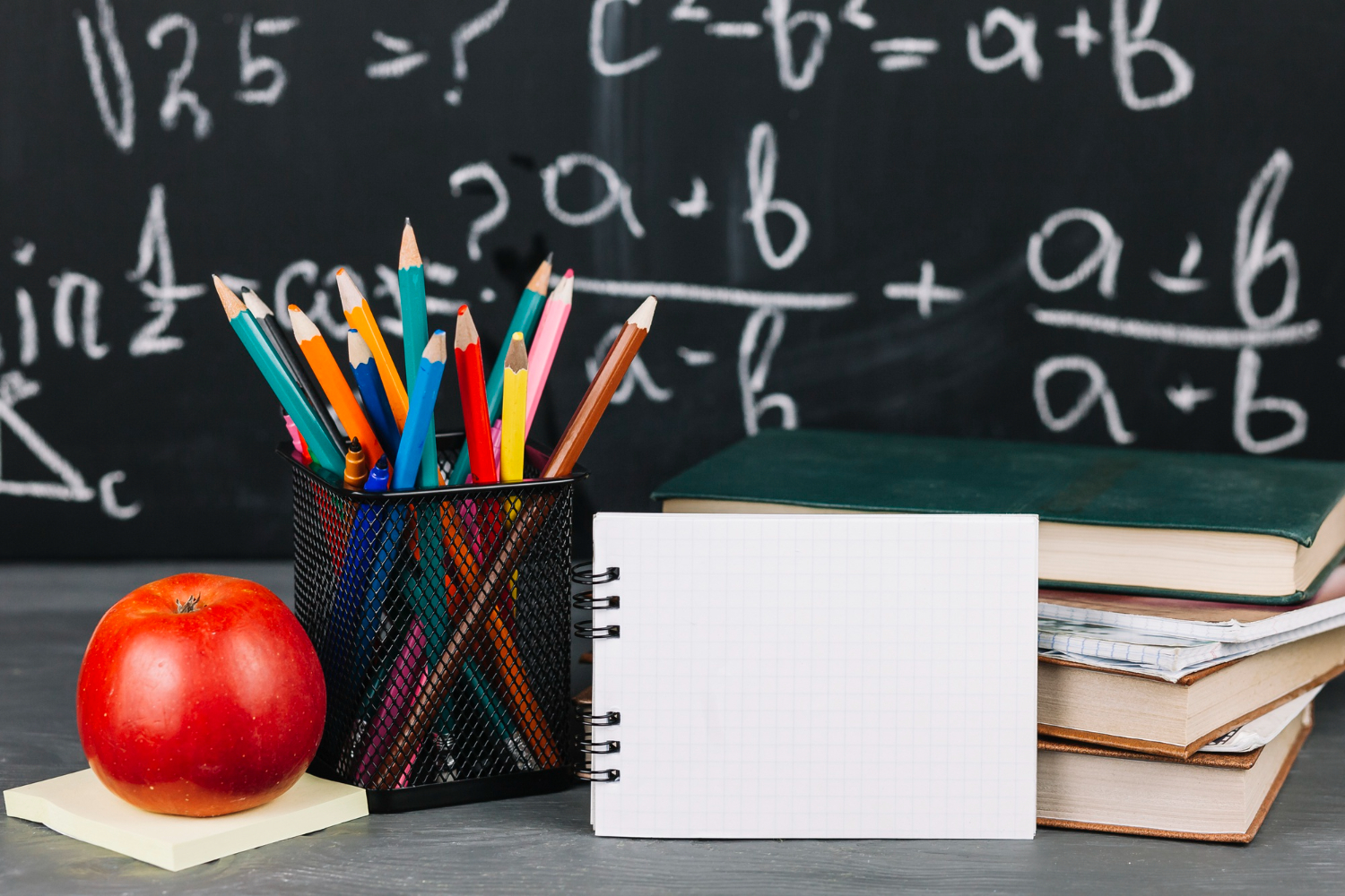 Biurko z jabłkiem, kolorowymi kredkami, otwartym zeszytem w kratkę i książkami, na tle tablicy z wzorami matematycznymi.